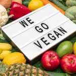 A lightbox sign reading "We Go Vegan" is surrounded by a variety of fruits and vegetables, including apples, bananas, lemons, artichokes, and cauliflower—an inviting snapshot to guide anyone eager to start a vegan life on a textured green surface.