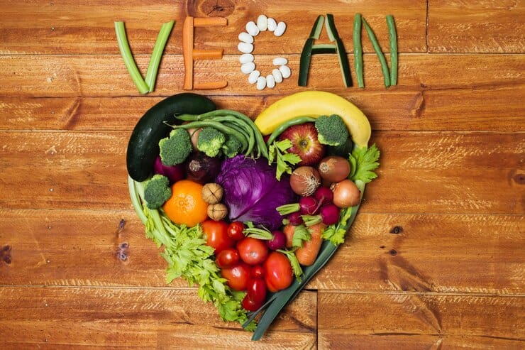 A heart shape made of various fruits and vegetables, including lettuce, tomatoes, broccoli, and bananas, is arranged on a wooden surface. Above it reads "Start Vegan," spelled out using different vegetables and beans.