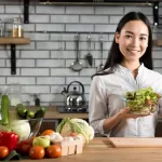 A woman in a kitchen gives a thumbs-up while holding a bowl of salad, embracing her vegan diet. The counter is filled with various vegetables like peppers, cucumbers, and tomatoes. A kettle and jars stand in the background against the tiled wall.
