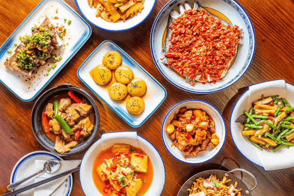 A vibrant top-view image of a wooden table filled with colorful Hunan cuisine dishes. The spread includes a variety of spicy and savory foods, such as Steamed Fish Head with Chili, stir-fried vegetables, braised pork, tofu in a rich chili sauce, golden steamed buns, and more. Each dish is served in traditional Chinese-style plates and bowls, garnished with fresh herbs, red chilies, and sesame seeds, highlighting the bold and flavorful characteristics of Hunan cuisine. 