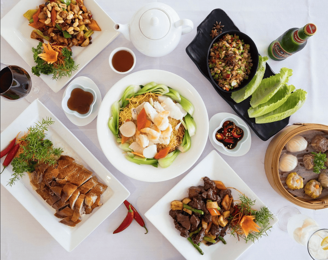 A top-view image showcasing an elegant presentation of Chinese cuisine on a white tablecloth. The spread includes various dishes such as roasted duck, stir-fried vegetables with nuts, seafood atop noodles, dim sum in a bamboo steamer, and lettuce wraps served with condiments. Accompanying the dishes are sauces, a white teapot, and beverages, creating a visually appealing and neatly arranged display. 