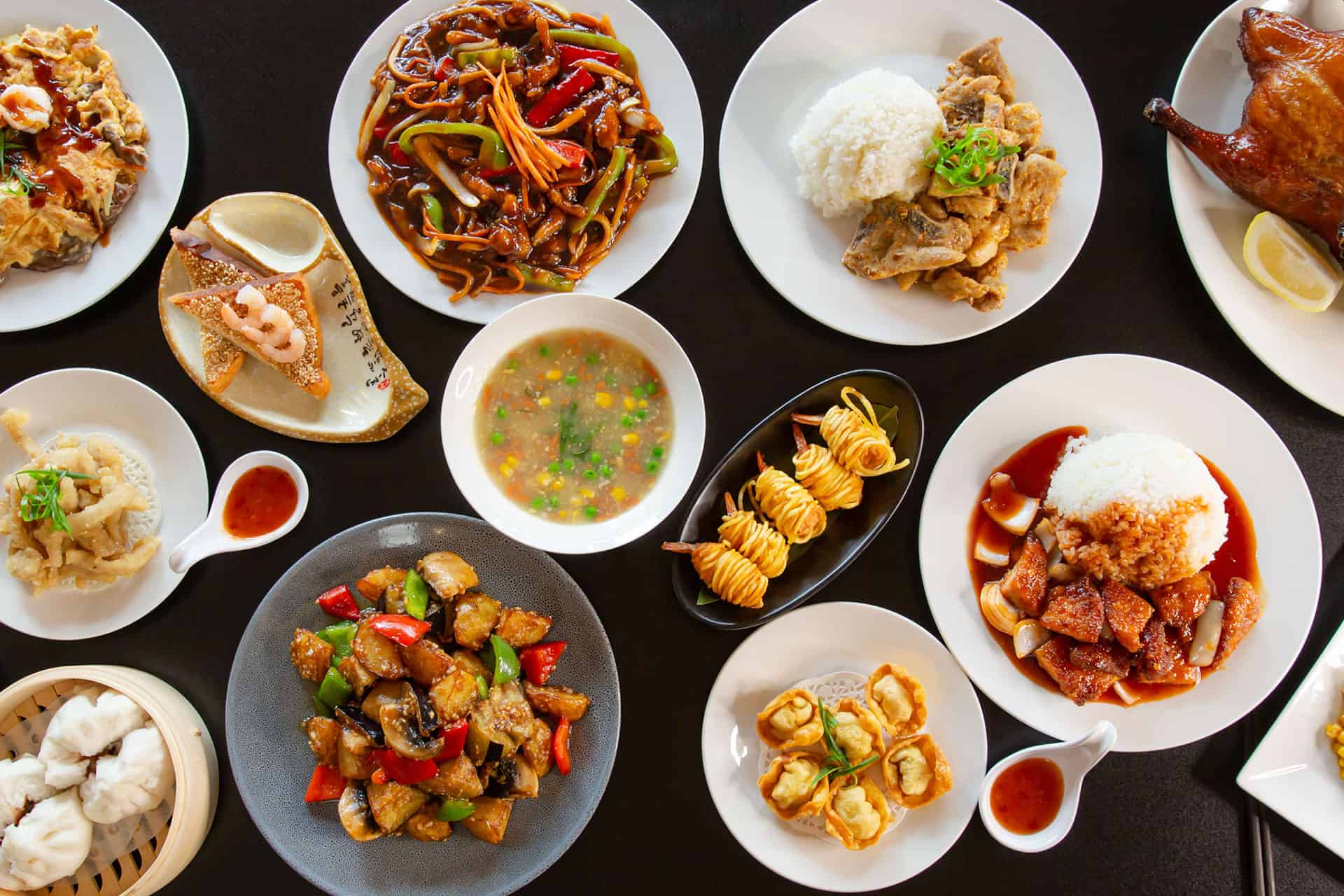 A top-view image showcasing a variety of Jiangsu cuisine dishes elegantly presented on white plates and a black table. The spread includes dishes like stir-fried vegetables, rice paired with savory meats, crispy fried appetizers, dumplings in a bamboo steamer, a colorful soup with peas and corn, and prawn rolls. Each dish is vibrant, emphasizing the fresh and mildly sweet flavors characteristic of Jiangsu cuisine, with an inviting and neatly arranged presentation. 