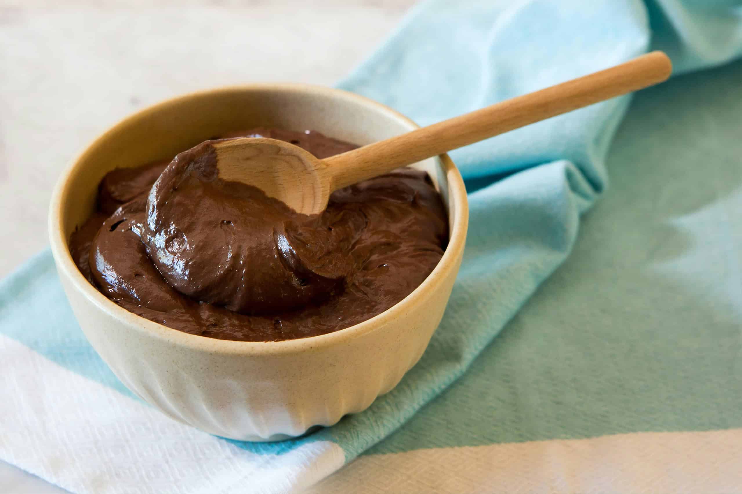 Close-up image of a bowl filled with smooth, rich vegan chocolate ganache. A wooden spoon rests in the ganache, highlighting its creamy texture. The bowl is placed on a light blue cloth, creating a soft and inviting presentation.