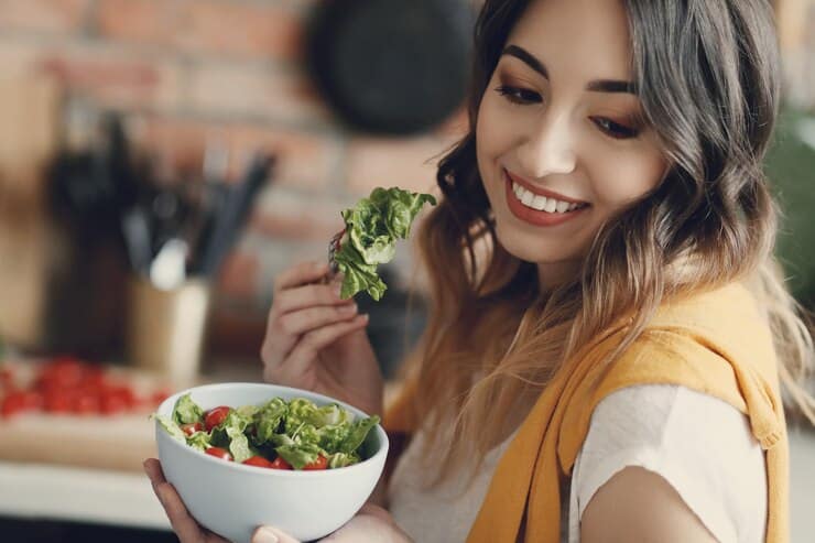 A woman smiles while holding a colorful bowl of fresh salad filled with various vegetables. The vibrant greens and reds are inviting.