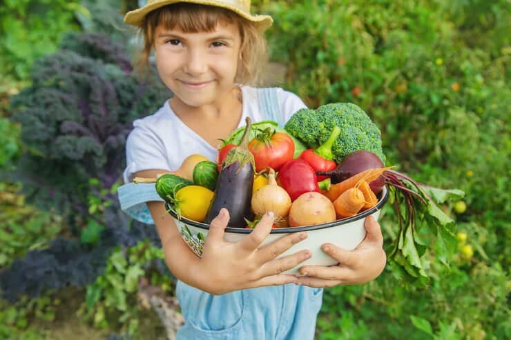 A smiling young girl wearing a straw hat and overalls holding a bowl filled with fresh vegetables, including broccoli, tomatoes, eggplant, potatoes, and carrots, standing in a vibrant garden.