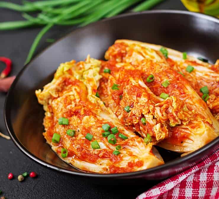 A black bowl filled with freshly prepared kimchi, featuring vibrant red chili paste coating Napa cabbage, garnished with chopped green onions. The dish is placed on a dark surface with scattered ingredients, including green onions and chili peppers in the background.