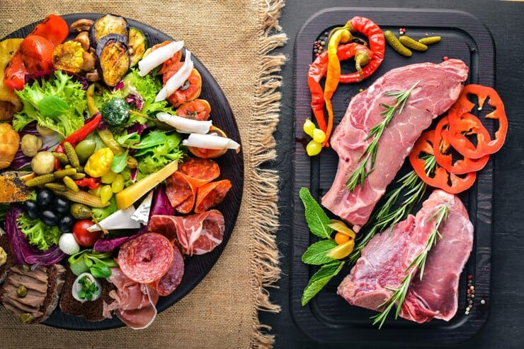 Two plates of food displayed side by side: on the left, a colorful assortment of fresh greens, pickled vegetables, cheeses, and cured meats arranged on a black platter; on the right, two raw steaks garnished with rosemary, surrounded by sliced tomatoes and chili peppers, presented on a dark serving tray.