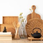 A minimalist kitchen setup featuring natural materials. The arrangement includes wooden cutting boards, woven placemats, a black cast iron teapot on a wooden stand, wooden utensils in a glass jar, and dried flowers in a glass vase. A small Buddha figurine sits on a stack of books, creating a calm and organized aesthetic.