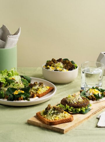 A plant-based meal arrangement featuring a wooden board with avocado toast, leafy greens, sprouts, and a plant-based burger patty. Surrounding it are a plate with a fresh green salad and a bowl of mixed salad with corn and leafy greens. A glass of water and a napkin holder complete the minimalist table setting on a light green tablecloth.  