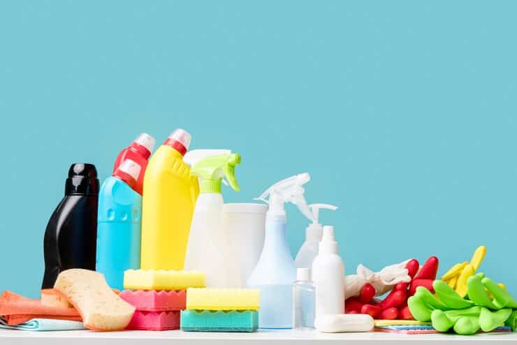 An assortment of colorful cleaning supplies arranged on a white surface against a light blue background. The items include spray bottles, detergent containers, sponges, scrubbing pads, gloves, and a cleaning cloth. The composition highlights a variety of cleaning tools and solutions.