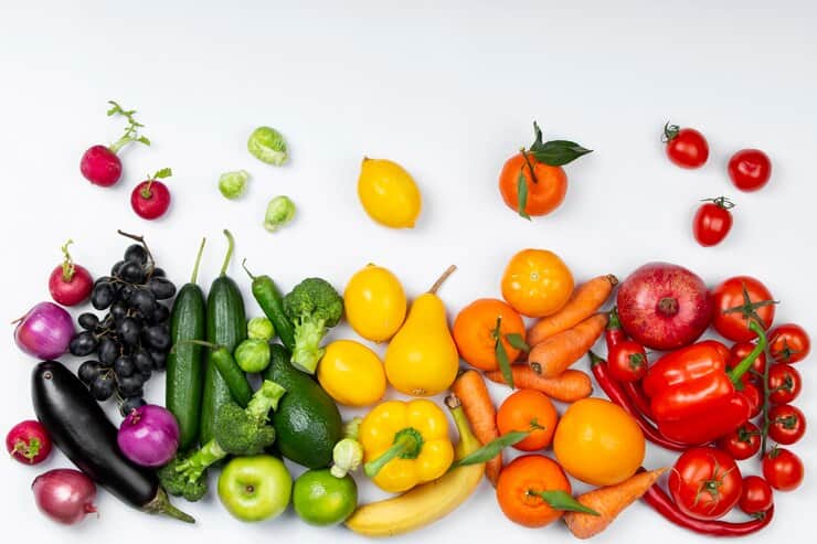 A colorful arrangement of fruits and vegetables organized by color, including radishes, eggplant, grapes, cucumbers, bell peppers, carrots, oranges, tomatoes, and more, displayed on a white background.