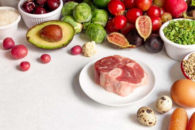 An assortment of fresh ingredients on a white surface, including a raw meat slice on a small white plate, surrounded by colorful produce like Brussels sprouts, figs, avocado, cherries, radishes, and peas, alongside bowls of grains, eggs, and quail eggs.