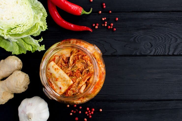 A top-down view of a jar filled with vibrant kimchi, placed on a dark wooden surface. Surrounding the jar are fresh ingredients for making kimchi, including Napa cabbage, red chili peppers, ginger, garlic, and scattered pink peppercorns, emphasizing the dish's fresh and traditional preparation.