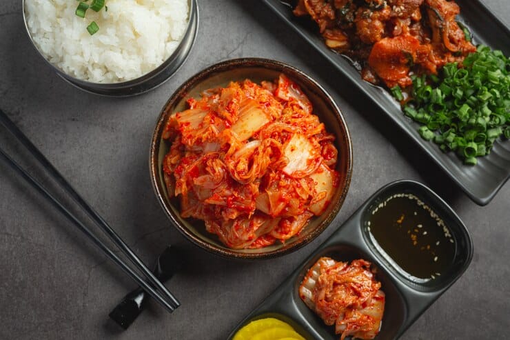 A bowl of vibrant kimchi as the centerpiece, surrounded by complementary dishes including steamed white rice, grilled meat, chopped green onions, soy dipping sauce, and side servings of pickled radish and additional kimchi. Black chopsticks rest on the table, completing the traditional Korean meal setup.