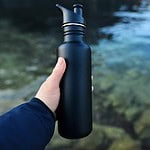 A person holding a sleek black reusable water bottle with a spout lid, standing near a clear natural water body with visible rocks underneath and a calm reflective surface in the background.