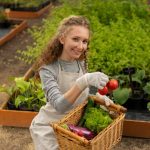 A smiling woman wearing gloves and an apron holding a fresh tomato and carrying a basket filled with vegetables, standing in a lush garden with raised planting beds.