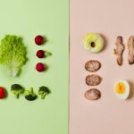 Two sets of food ingredients arranged side by side on a split green and pink background: on the green side, fresh vegetables including lettuce, radishes, broccoli, cherry tomatoes, and parsley; on the pink side, cooked items such as bacon strips, sausage slices, a halved hard-boiled egg, and a doughnut.