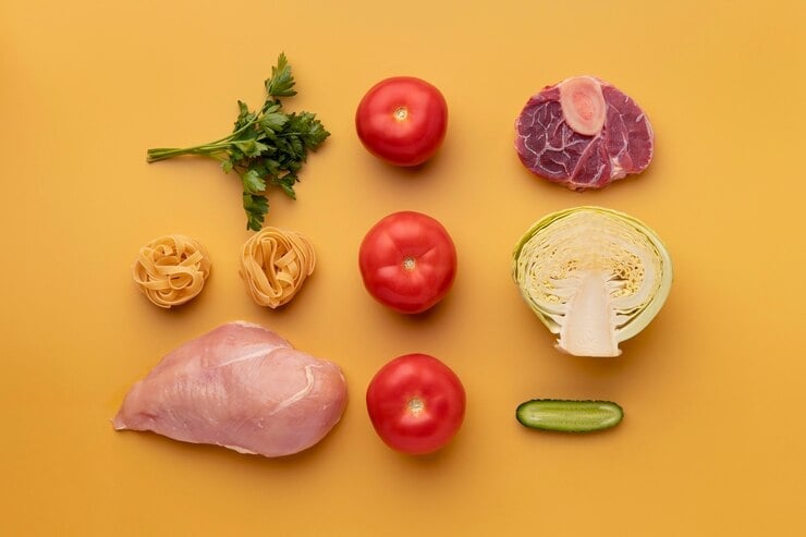 A neatly arranged selection of fresh ingredients on a yellow background, including raw chicken breast, a beef cut with bone, whole tomatoes, parsley, a halved cabbage, a cucumber slice, and two nests of pasta.