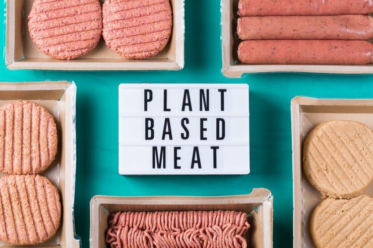 The image shows several packages of plant-based meat alternatives, including patties, sausages, and ground meat, arranged around a lightbox sign displaying the text "PLANT BASED MEAT" on a turquoise background.  