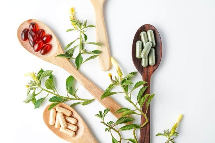 Wooden spoons holding different types of dietary supplements, including red soft gels, beige capsules, and green capsules, arranged on a white background. Fresh green leaves and small yellow flowers are scattered around, emphasizing a natural and herbal theme.