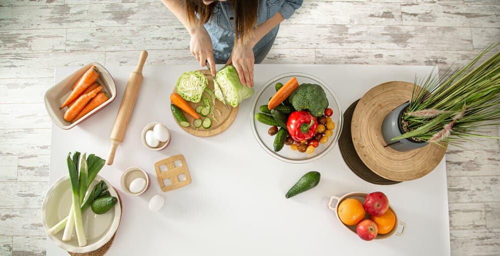 A woman is chopping colorful vegetables on a white table, viewed from above, showcasing her culinary preparation.