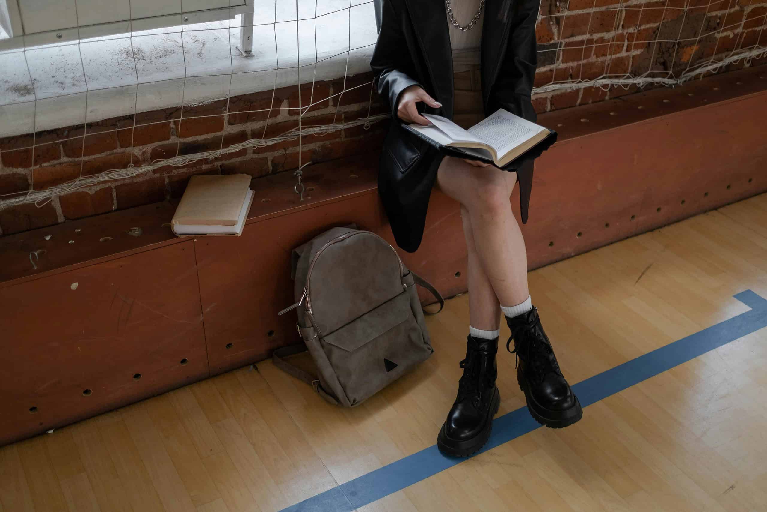 A woman in a vegan leather jacket, bag, and boots sits on a bench, engrossed in reading a book.