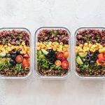 The image shows three meal prep containers filled with healthy, colorful ingredients. Each container includes purple rice, corn, cherry tomatoes, cucumber, black olives, green onions, and lentils, neatly arranged in rows, showcasing a vibrant and balanced meal.