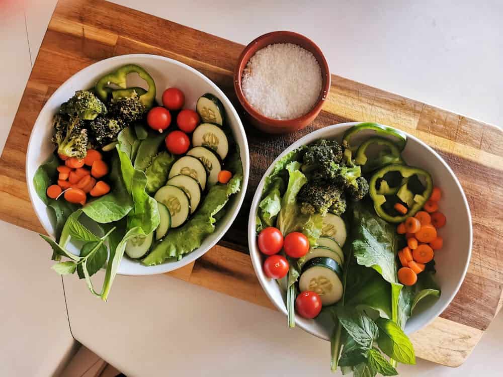 Two colorful bowls filled with fresh vegetables sit on a wooden cutting board, ready for meal preparation. The vibrant colors enhance the kitchen scene.