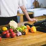 A person in a white shirt stands in a kitchen behind a wooden counter filled with fresh vegetables and produce. The assortment includes tomatoes, cucumbers, garlic, bell peppers in green, orange, and yellow, cauliflower, eggplants, and a lemon. The organized setup suggests meal preparation with vibrant, colorful ingredients.