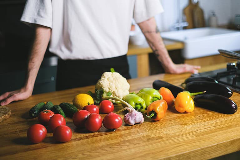 A person in a white shirt stands in a kitchen behind a wooden counter filled with fresh vegetables and produce. The assortment includes tomatoes, cucumbers, garlic, bell peppers in green, orange, and yellow, cauliflower, eggplants, and a lemon. The organized setup suggests meal preparation with vibrant, colorful ingredients.