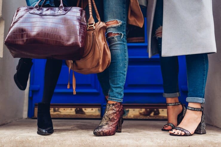 Three women in jeans and boots stand on a sidewalk, showcasing the latest vegan leather bags and boots fashion trends for 2025.