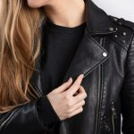 A young female wearing a vegan leather jacket standing against white wall.