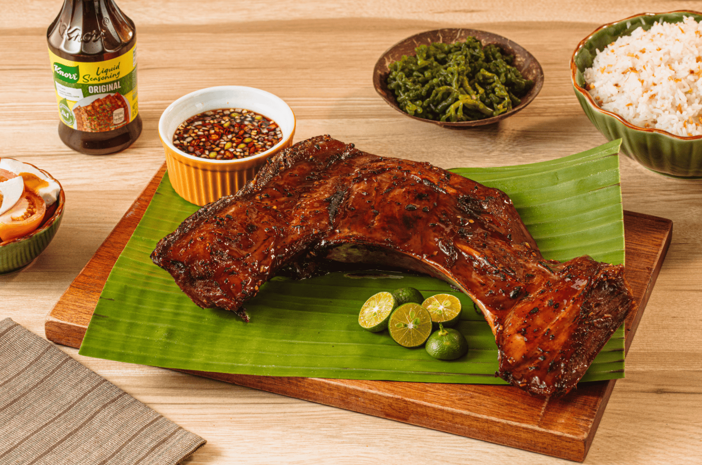 A succulent slab of grilled pork ribs glazed with a rich, caramelized marinade, served on a banana leaf for an authentic Filipino touch. Accompanying the ribs are a dipping sauce of soy sauce with chili and onions, steamed rice, fresh vegetables, and calamansi for a zesty flavor boost. The rustic wooden board and vibrant condiments complete the appetizing presentation of this Filipino classic. 