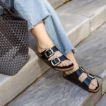 A person sitting on gray stone steps, wearing black double-strap sandals with gold buckles and frayed-hem light blue jeans. A woven black tote bag is placed beside them, complementing the casual yet stylish outfit.