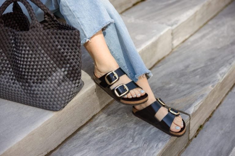 A person sitting on gray stone steps, wearing black double-strap sandals with gold buckles and frayed-hem light blue jeans. A woven black tote bag is placed beside them, complementing the casual yet stylish outfit.