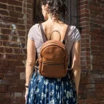 A woman wearing a brown leather backpack stands with her back facing the camera against a rustic brick wall. She is dressed in a striped short-sleeve top and a blue floral skirt, with her dark, curly hair tied up in a loose ponytail.