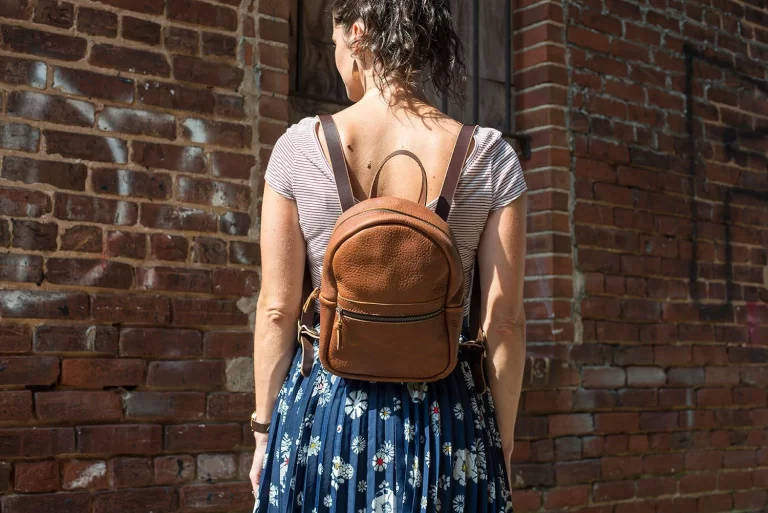 A woman wearing a brown leather backpack stands with her back facing the camera against a rustic brick wall. She is dressed in a striped short-sleeve top and a blue floral skirt, with her dark, curly hair tied up in a loose ponytail.