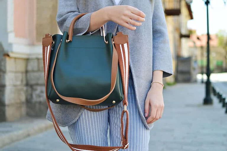 A fashionable woman carrying a dark green handbag with brown leather handles and a striped shoulder strap. She is wearing a light gray coat over striped blue and white pants, standing on a European-style street with stone buildings and lampposts in the background.