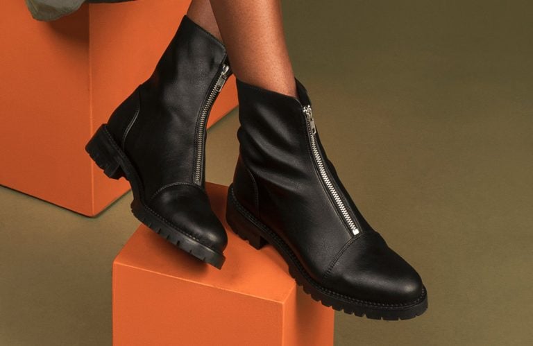 A pair of black leather ankle boots with front zippers and rugged soles, worn by a model posing with one foot resting on an orange geometric block. The boots have a sleek yet edgy design, contrasting with the warm-toned background.