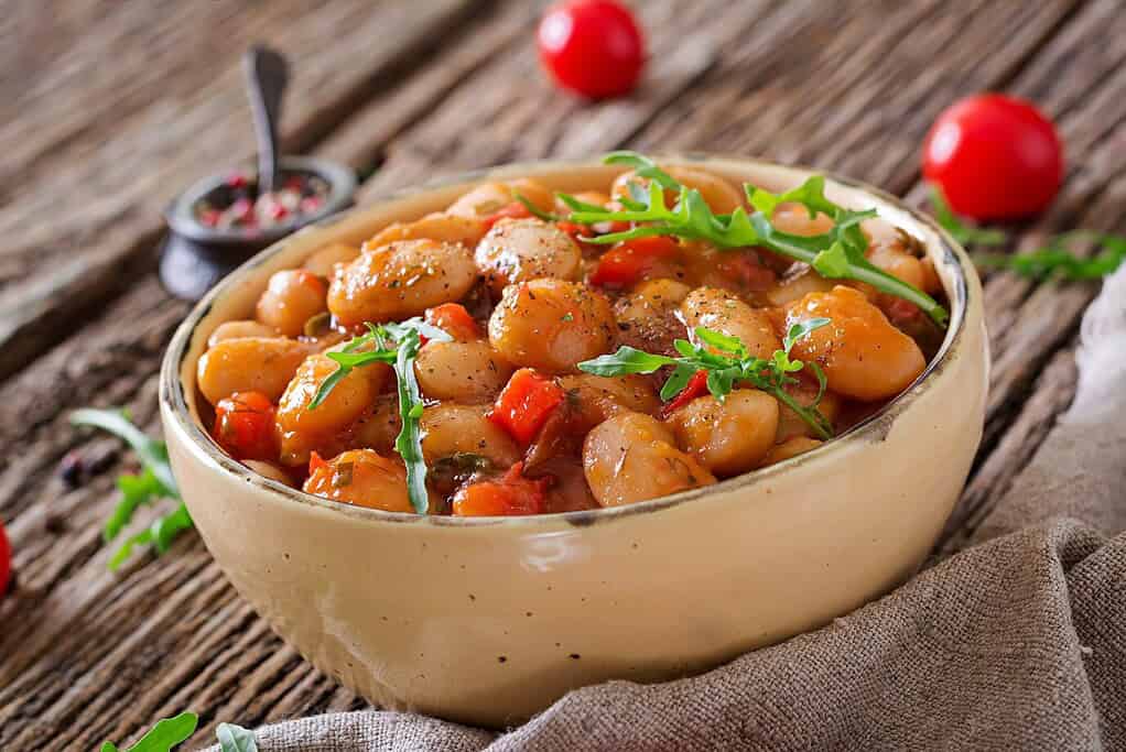 A rustic bowl filled with butter beans in tomato-based sauce, garnished with fresh herbs and surrounded by cherry tomatoes on a wooden table.
