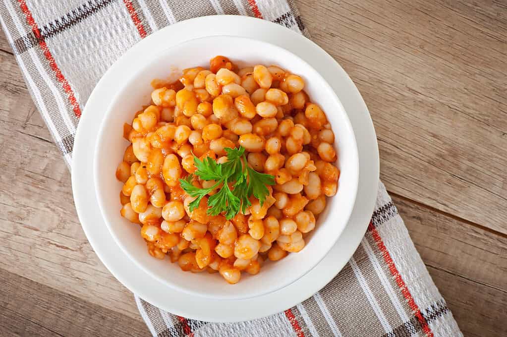 Butter beans served in a white bowl with rich tomato sauce and garnished with fresh parsley on a rustic wooden table.